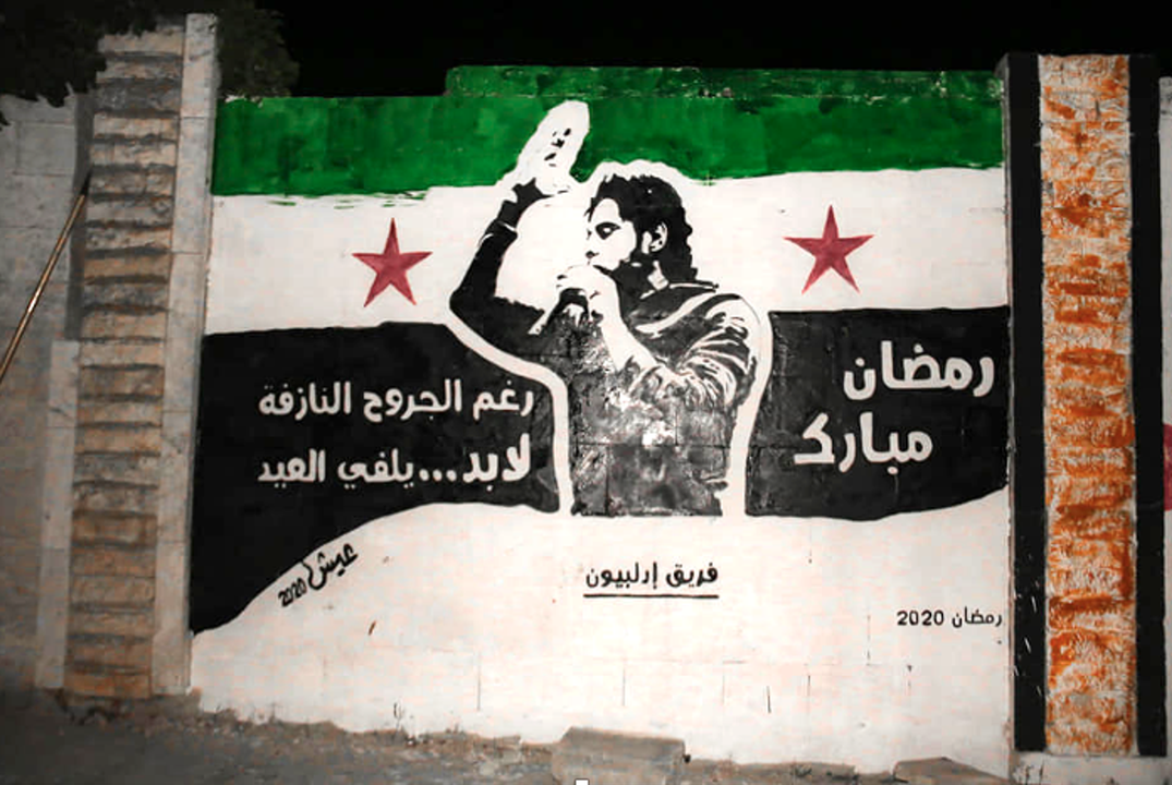 Figure 4. Mural painted in 2020 by the Aish Campaign (Live Campaign), a grassroot political art collective in Kafr Nabl, Idlib province, depicts Abdul Baset al-Sarout against the backdrop of the Syrian revolu-tionary flag. The message reads: “Blessed Ramadan. Despite the bleeding wounds, inevitably. . . Eid is coming” (“Ramaḍān mubārak. Raghma al-jurūḥ al-nāzifah,lābud. . . yalffī al-ʿīd”; artwork and photo credit: Aish Campaign [حملة عيش] 2020, with kind permission).