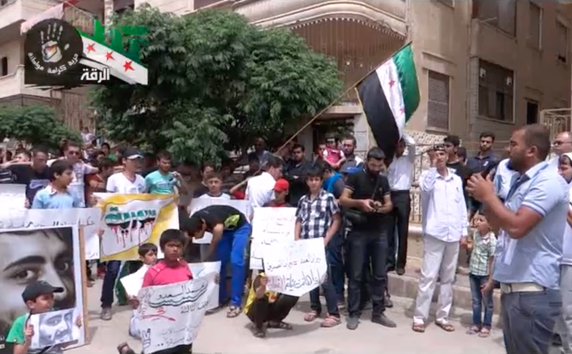 Figure 5. Ismail Gadaan (on the right) sings “Bi-ḥuzn wa-al-rīḥ taṣfar min warā al-bībān” during a protest outside the family home of the deceased Mohammed al-Saʿdo (his photo is visible on the left) in al-Thakana, Raqqa, in May 2013 (still by the author from a YouTube video uploaded by Raqqa Rev 2013).